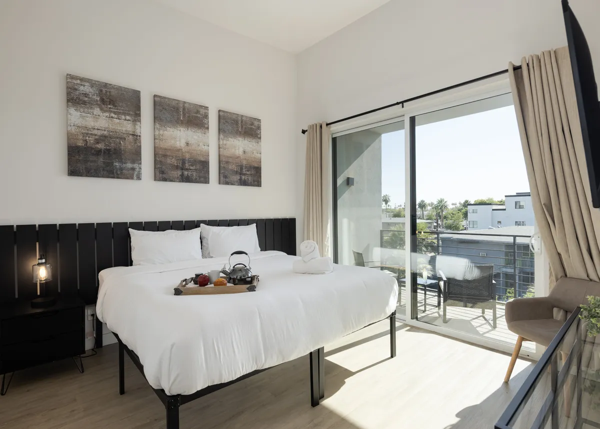 This image displays a serene bedroom with a king-size bed, tasteful artwork on the wall, a private balcony visible through glass doors, and a tray with refreshments on the bed. Sunlight filters softly through the curtains, creating a cozy and inviting atmosphere ideal for relaxation.