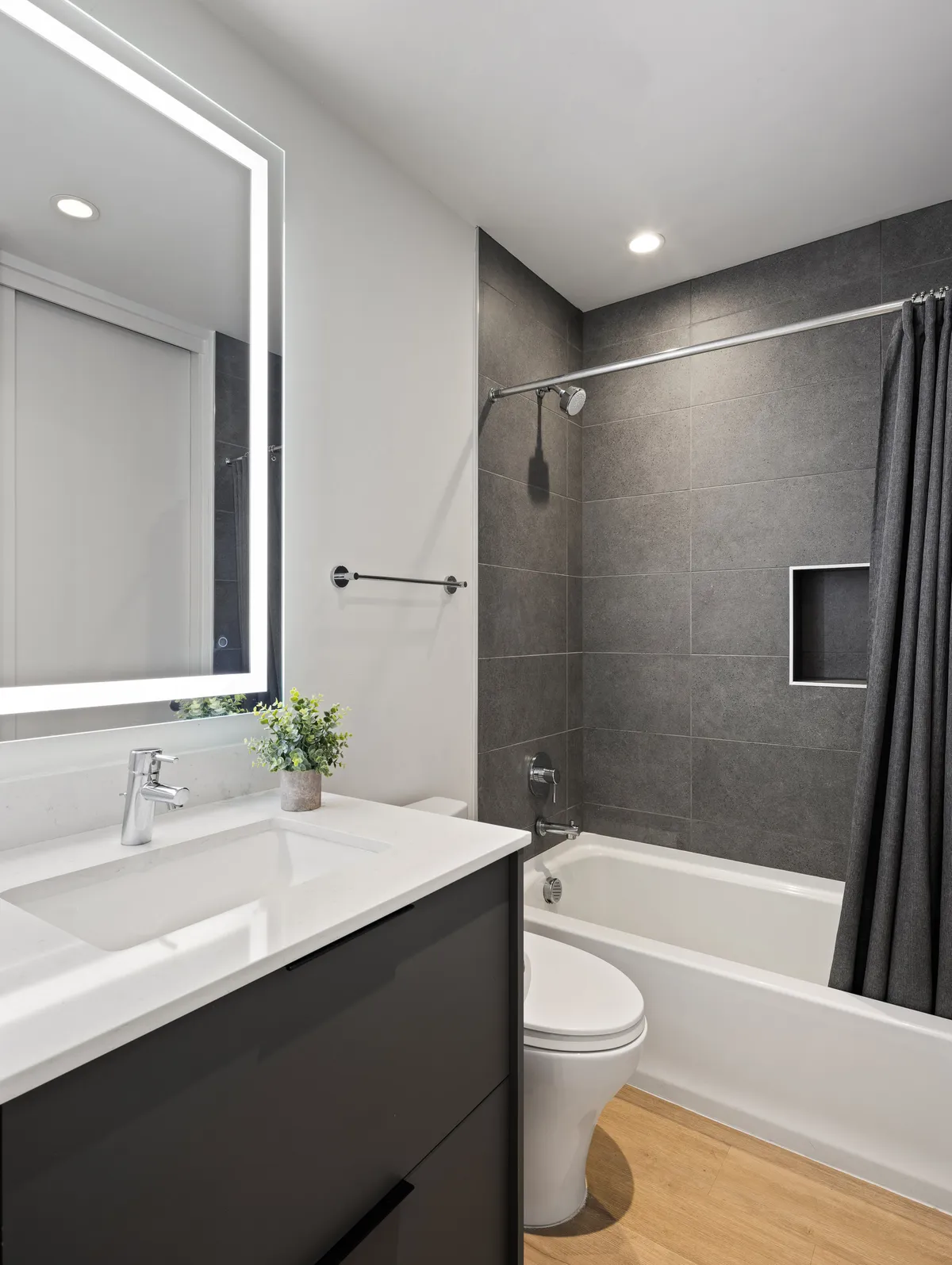 A calm, modern ensuite bathroom with a vanity, sink, backlit mirror, bathtub, shower curtain, and contemporary wall tiles. The space feels soothing and perfect for relaxation after a long day.