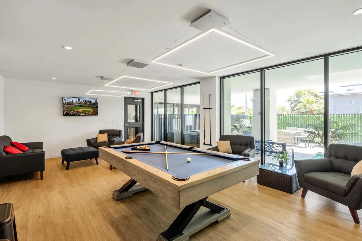 A contemporary community room featuring a pool table at the center, surrounded by plush sofas and a large TV. Sunlight pours in through expansive windows, creating an inviting, casual atmosphere ideal for socializing and friendly competition.