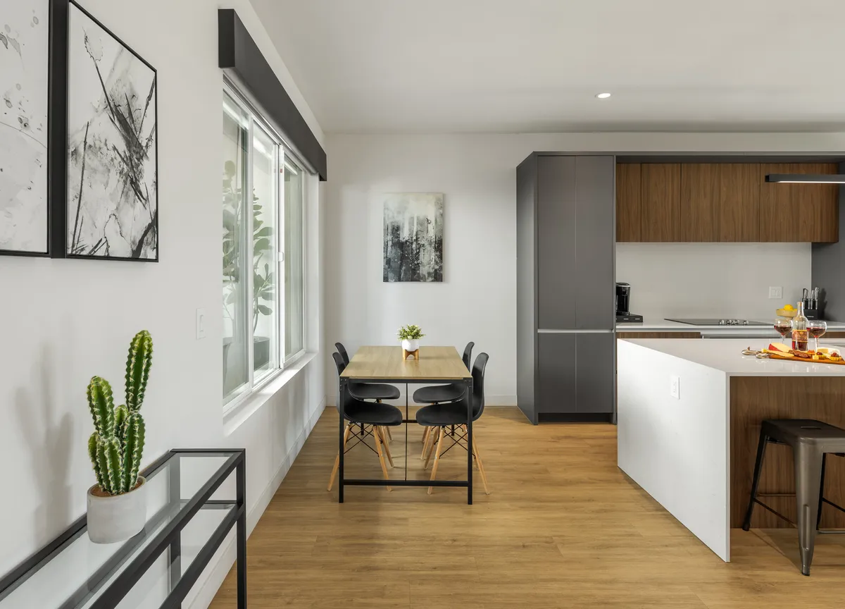 A bright, inviting kitchen and dining area featuring a contemporary dining table, cactus plant centerpiece, and sleek kitchen in the background. Artwork and sunlight create a welcoming, flexible space for meals or gatherings.
