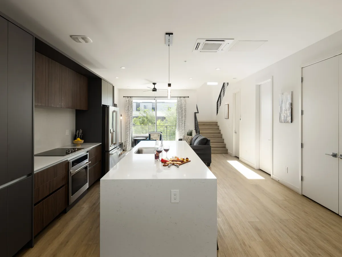 A welcoming interior shot showing the flow from the kitchen island to the dining area, with a staircase and balcony view in the background. The bright, modern decor and open sight lines encourage both connection and retreat, reflecting the social sanctuary theme.
