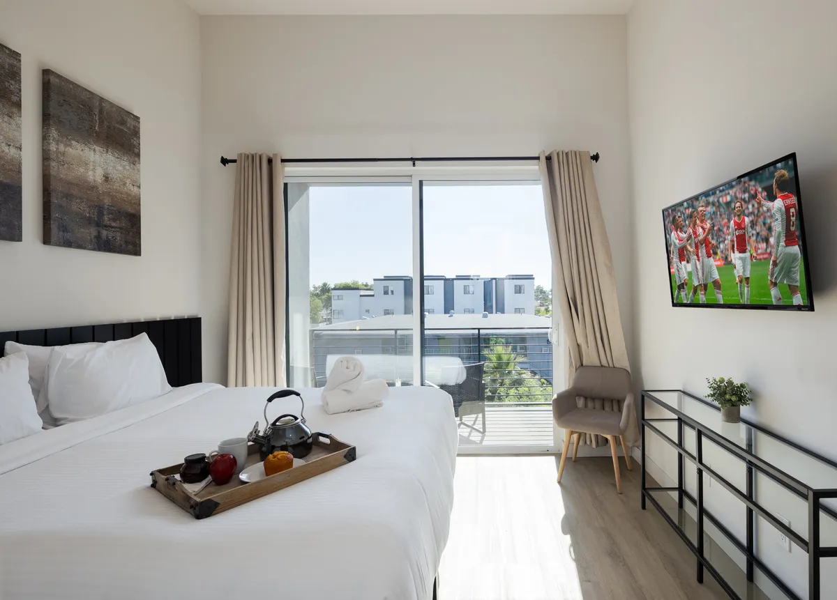 The scene features a comfortable bed with crisp white linens, a tray with tea and snacks, a television displaying a sporting event, and a large window bathing the space in natural light. The proximity to the balcony is suggested, enhancing the sense of indoor-outdoor living.