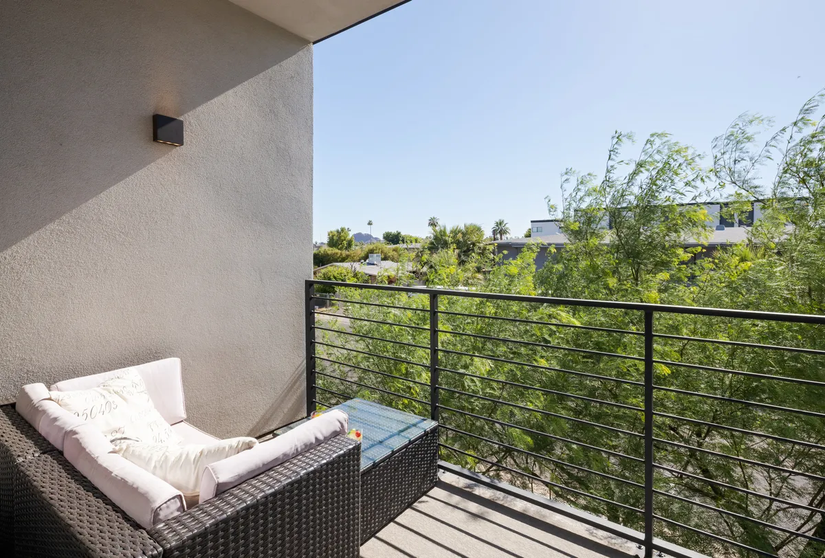 A private balcony with a wicker couch adorned by a decorative pillow, framed by a balcony railing and overlooking lush greenery. The scene is calm, with soft lighting suggesting early evening or morning, emphasizing a personal outdoor retreat for relaxation or casual work breaks.