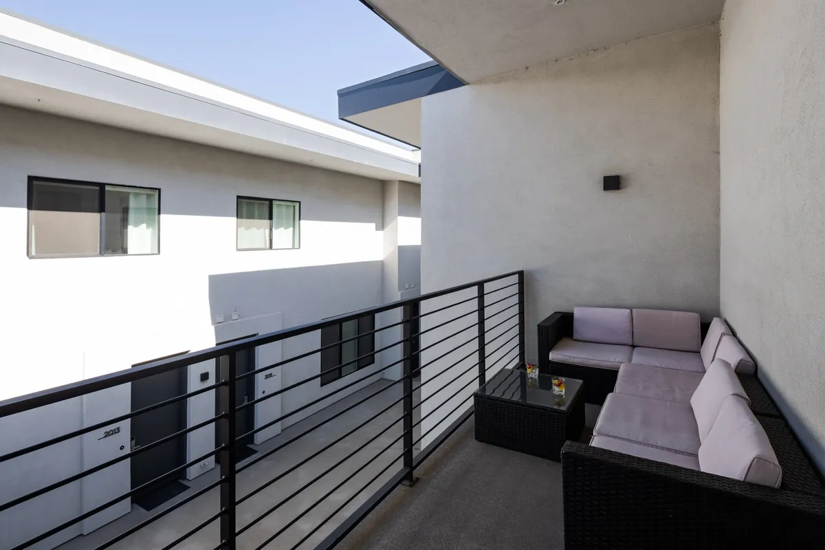 A casually arranged balcony seating area featuring a small table with drinks, comfortable outdoor chairs, and a view of neighboring modern buildings. The area is bathed in morning light, suggesting a pleasant spot for coffee, socializing, or evening relaxation.