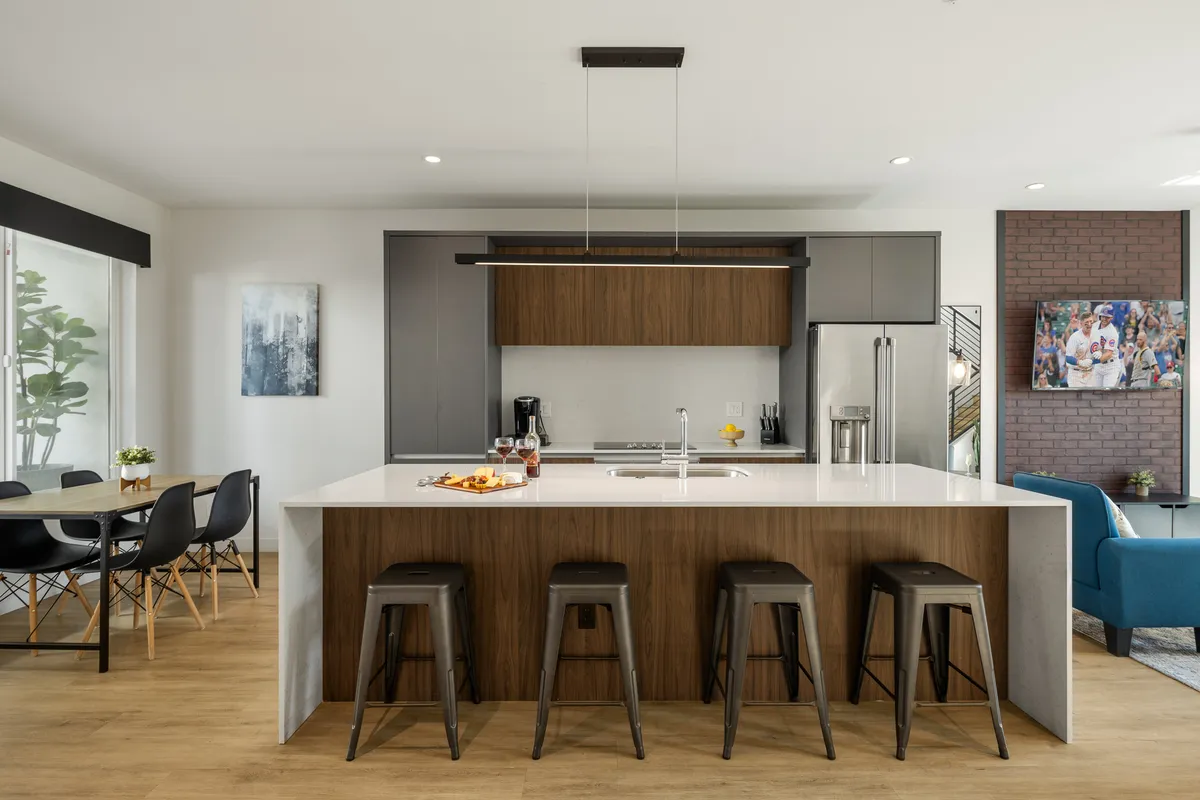 This image presents a bright, open-concept living space that seamlessly integrates the kitchen, dining table, and a comfortable seating area. Visible are contemporary furnishings, artwork on the walls, and a warm, inviting atmosphere ideal for both entertaining and quiet relaxation. The layout highlights both the culinary and social aspects of the space.