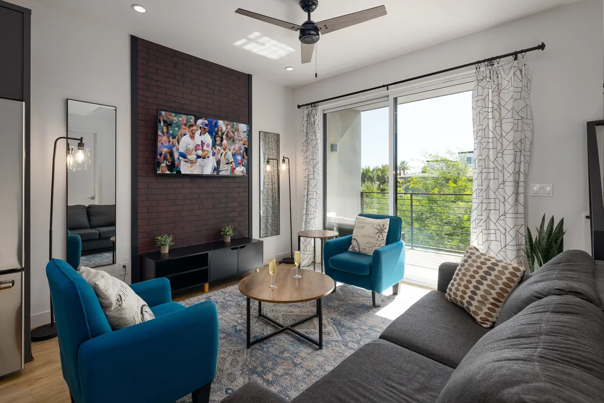 The image showcases a bright, inviting living area with plush blue armchairs facing a wall-mounted TV, a stylish wooden coffee table in the center, and large windows flooding the space with natural light. The setup suggests a perfect environment for relaxing, watching sports, or gathering with friends, with glimpses of Scottsdale's sunshine just outside.